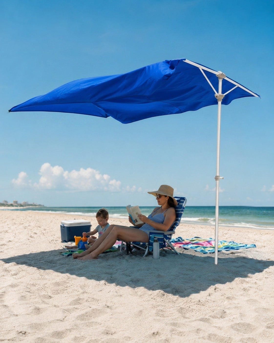 A large blue beach umbrella with a white pole is set up on a sandy beach, shading a woman and child sitting in lounge chairs near the ocean.