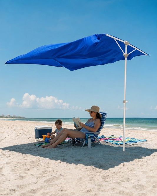 A large blue beach umbrella with a white pole is set up on a sandy beach, shading a woman and child sitting in lounge chairs near the ocean.