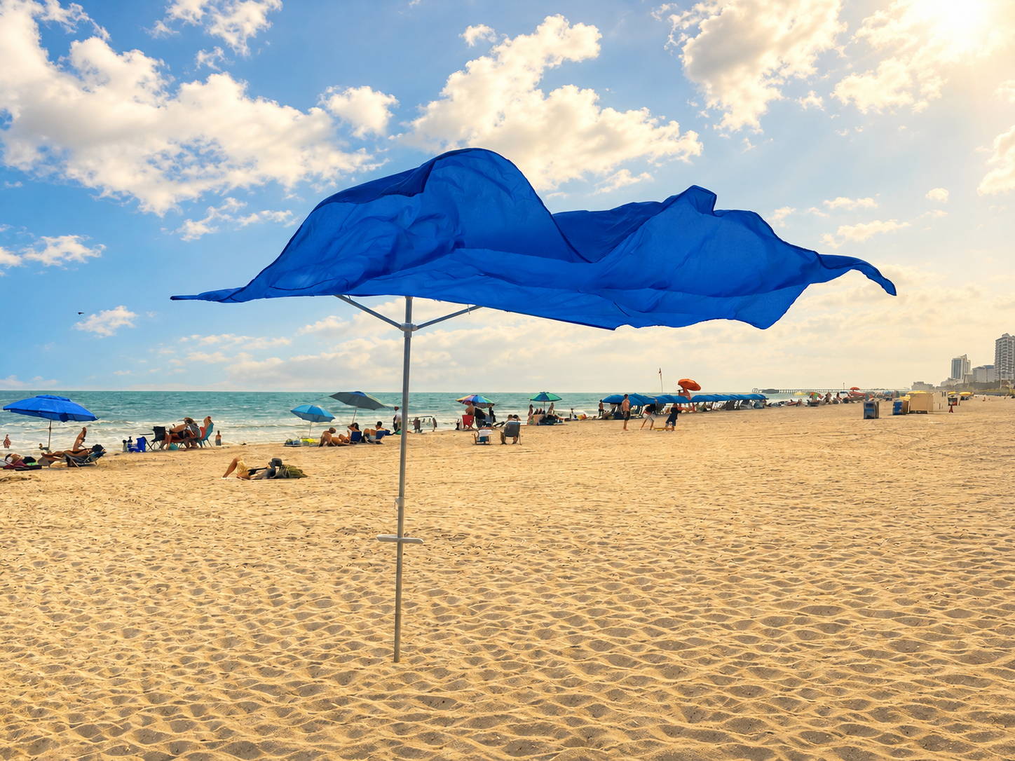 A large blue beach umbrella is shown on a sandy beach with the ocean, sky, and clouds in the background.