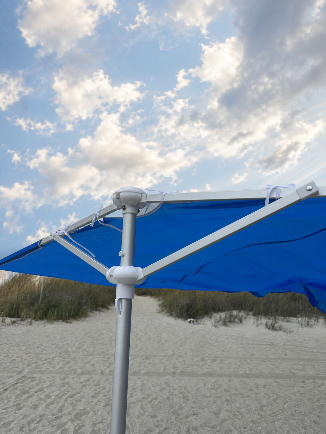 A close-up outdoor shot of a blue patio umbrella canopy attached to a white metal frame, with a sandy beach and cloudy sky in the background.